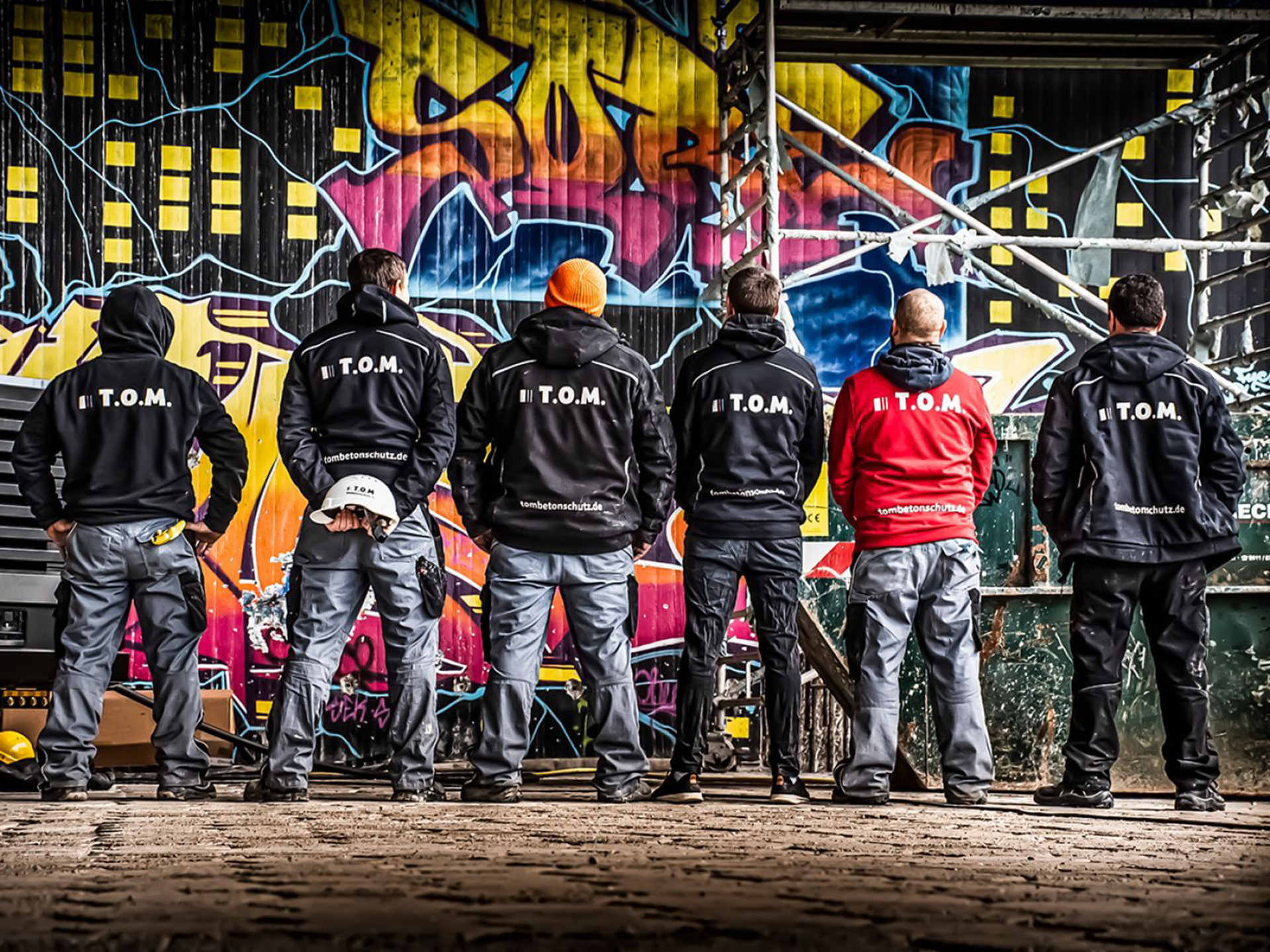 Gruppenaufnahme des Teams von „T.O.M.“: Sechs Männer stehen mit dem Rücken zur Kamera vor einer bunten Graffitiwand, daneben ein Baugerüst und ein Container.