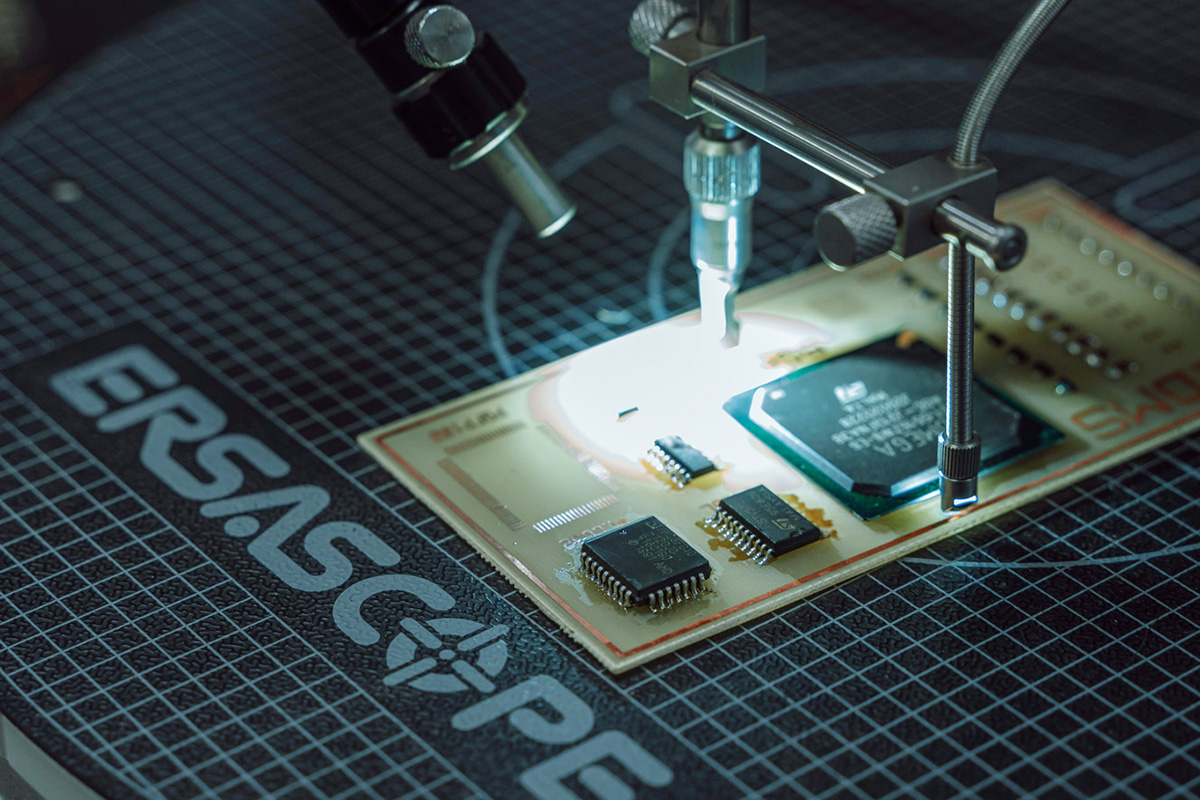 Platine mit mehreren Mikrochips, darüber ein Werkzeug an einem Haltearm, das die Leiterplatte im hellen Lichtkegel bearbeitet.
